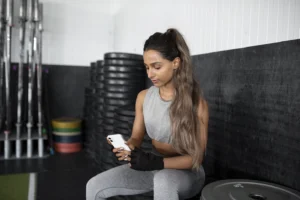 Mulher na academia olha o celular com expressão de dúvida ao escolher um personal trainer no app PersonalGO.